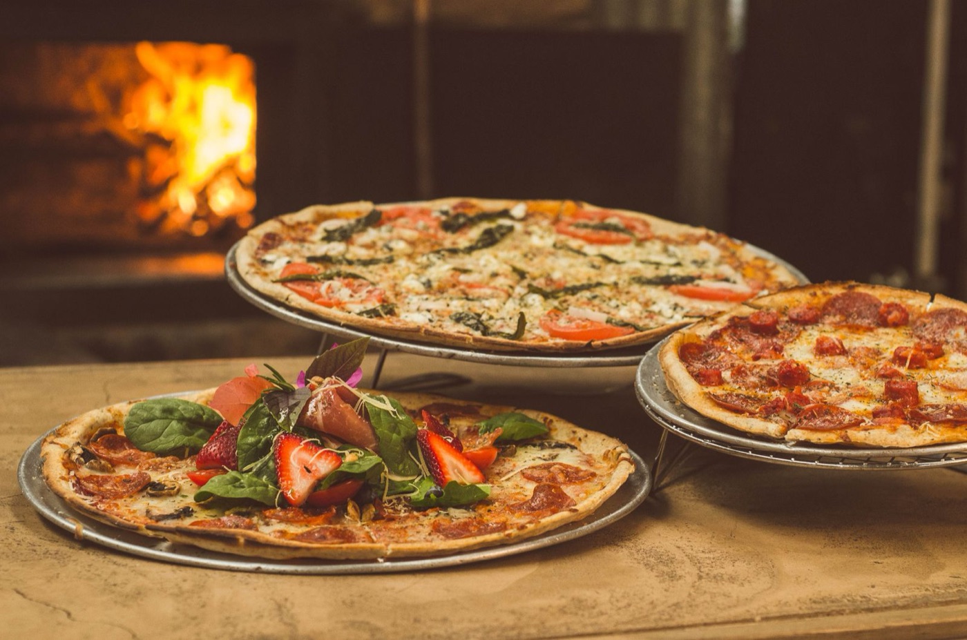 Three pizzas with fresh toppings near a wood-fired oven